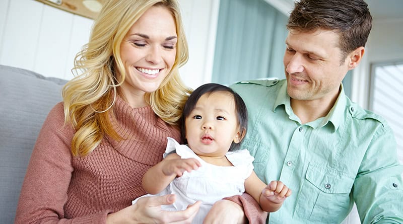 Un couple européen souriant assis sur un canape, la femme a une petite fille asiatique sur les genoux.