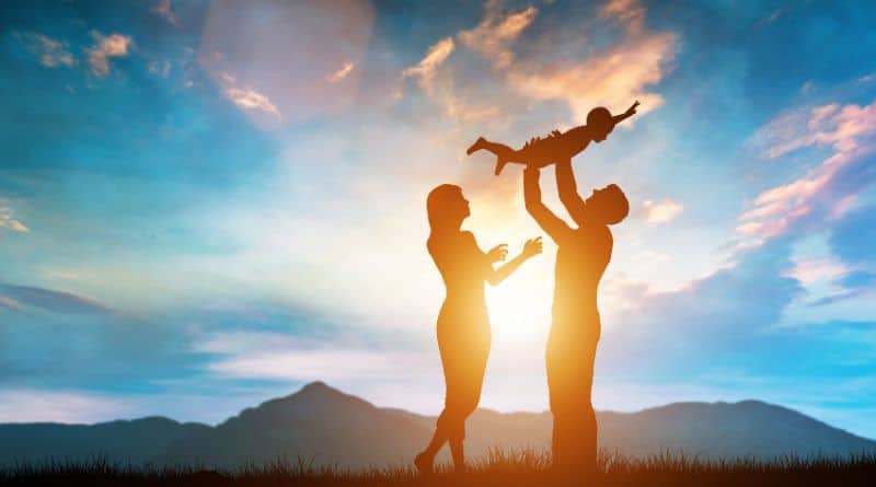 Un couple dans la nature avec un enfant dans les bras de l'homme en contre jour avec un couché de soleil des montagnes en fond.