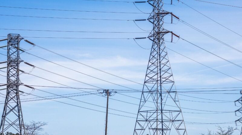 Dans la campagne, au premier plan 2 pylones électriques de tres haute tension, au second plan un pylone haute tension sur ciel bleu